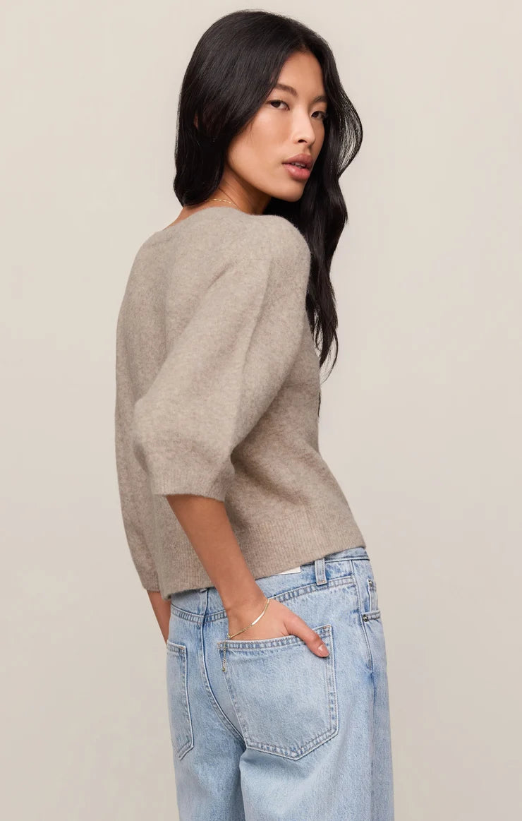 A woman wearing a light taupe knit sweater and light blue jeans, photographed from the back-three-quarter angle against a plain light beige studio background.