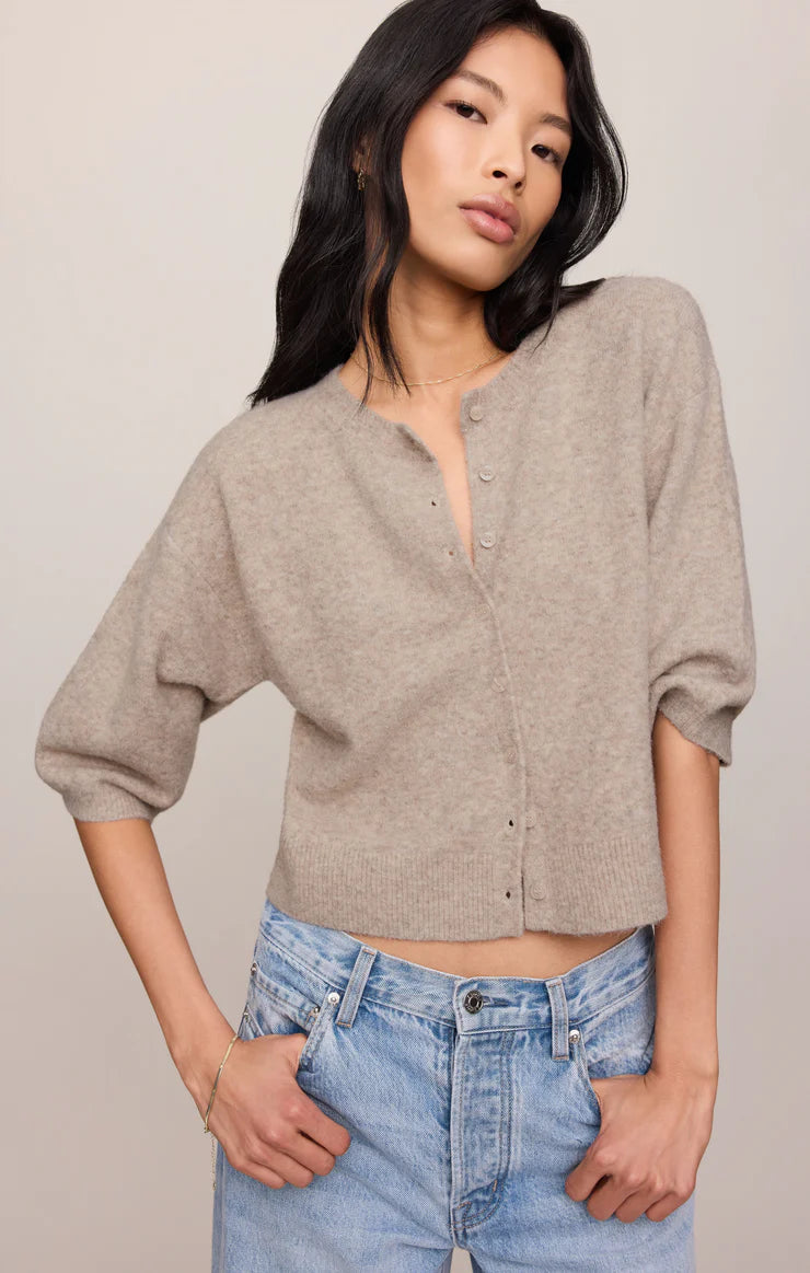 Woman wearing a beige button-up cardigan and blue jeans against a plain background