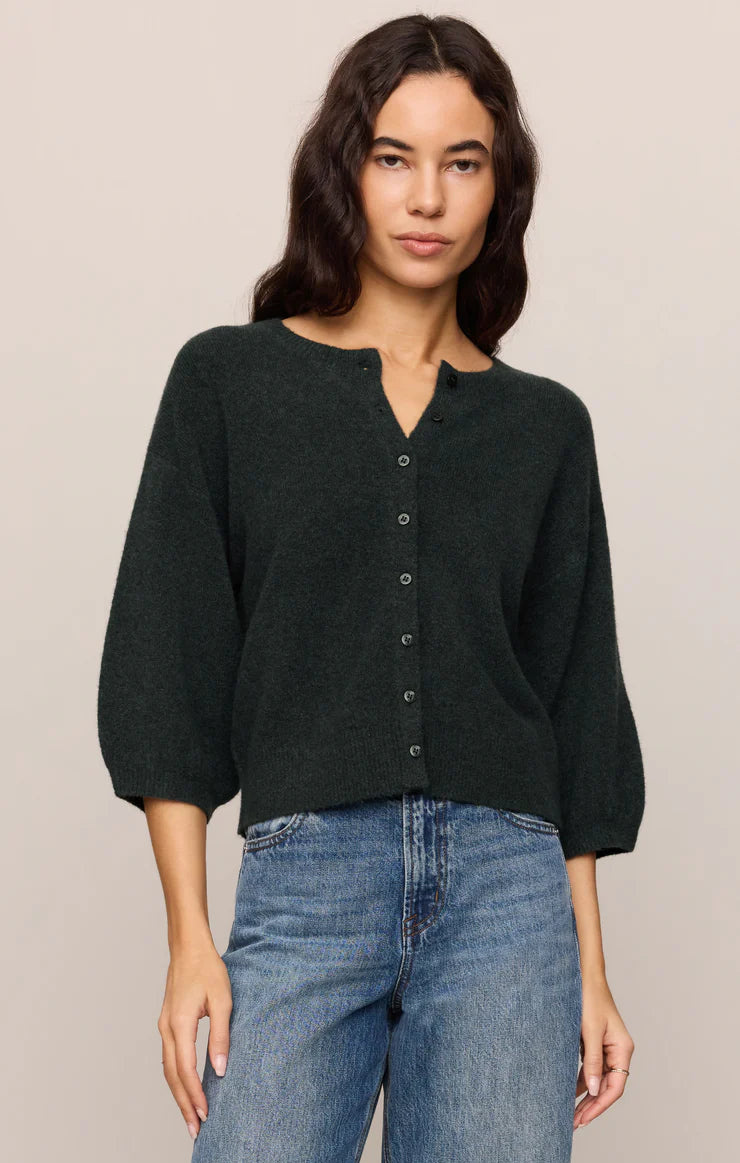 A woman wearing a dark forest green knit cardigan with a buttoned split neckline and three-quarter sleeves, paired with blue jeans, against a solid light beige background.