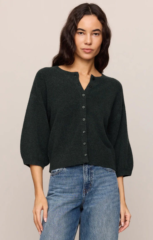 A woman wearing a dark forest green knit cardigan with a buttoned split neckline and three-quarter sleeves, paired with blue jeans, against a solid light beige background.