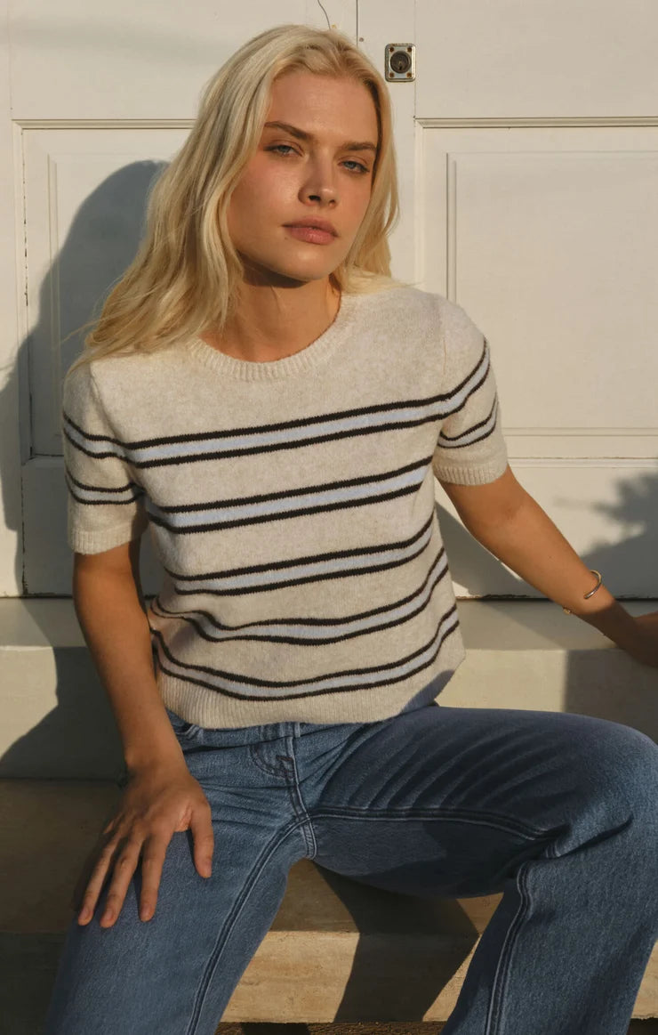 A blonde woman sitting outdoors against a white paneled wall, wearing a cream knit sweater with thin black horizontal stripes and blue jeans.