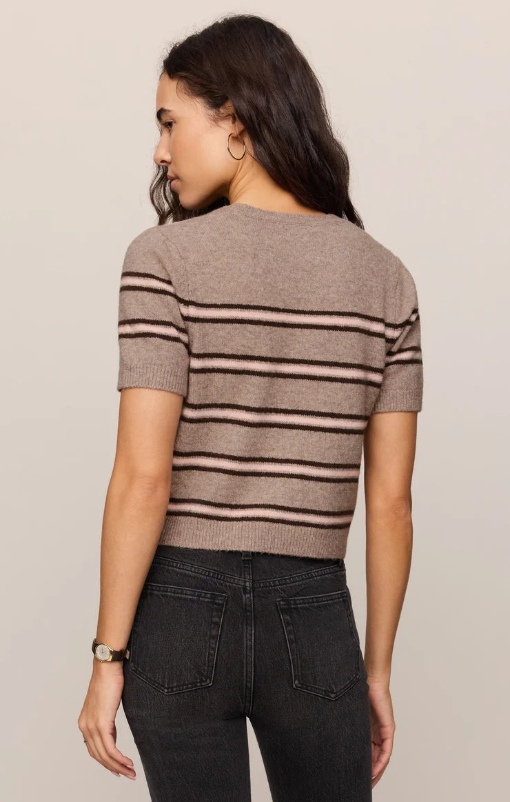 A woman shown from the back wearing a short-sleeve knit top with horizontal taupe and black stripes, paired with dark gray jeans, against a plain light beige studio background.