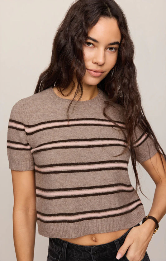 A woman wearing a short-sleeve knit crop top with horizontal stripes in taupe, black, and light pink, paired with dark jeans against a plain beige background.